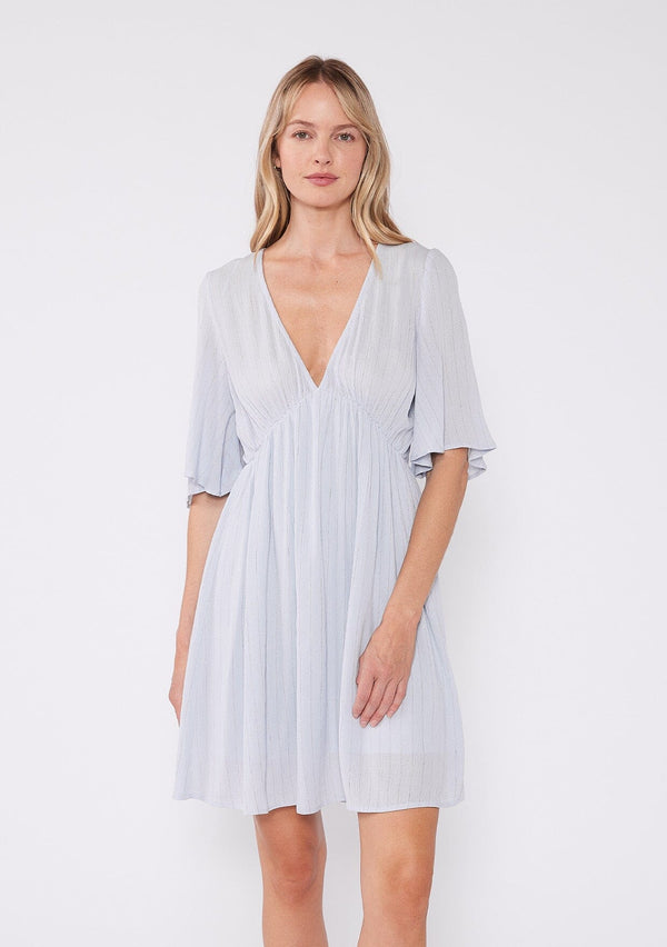 [Color: Periwinkle] A front facing image of a blonde model wearing a classic flowy mini dress in light blue with a gold metallic thread detail. With short half length sleeves, a v neckline, and an empire waist.