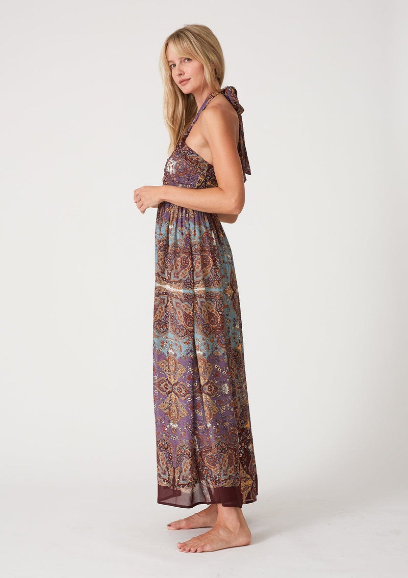 [Color: Raisin/Teal] A side facing image of a blonde model wearing a bohemian halter maxi dress in a multi colored purple and teal floral print. With a tie neckline, a slim fit bodice with a smocked elastic back, and a long flowy skirt.