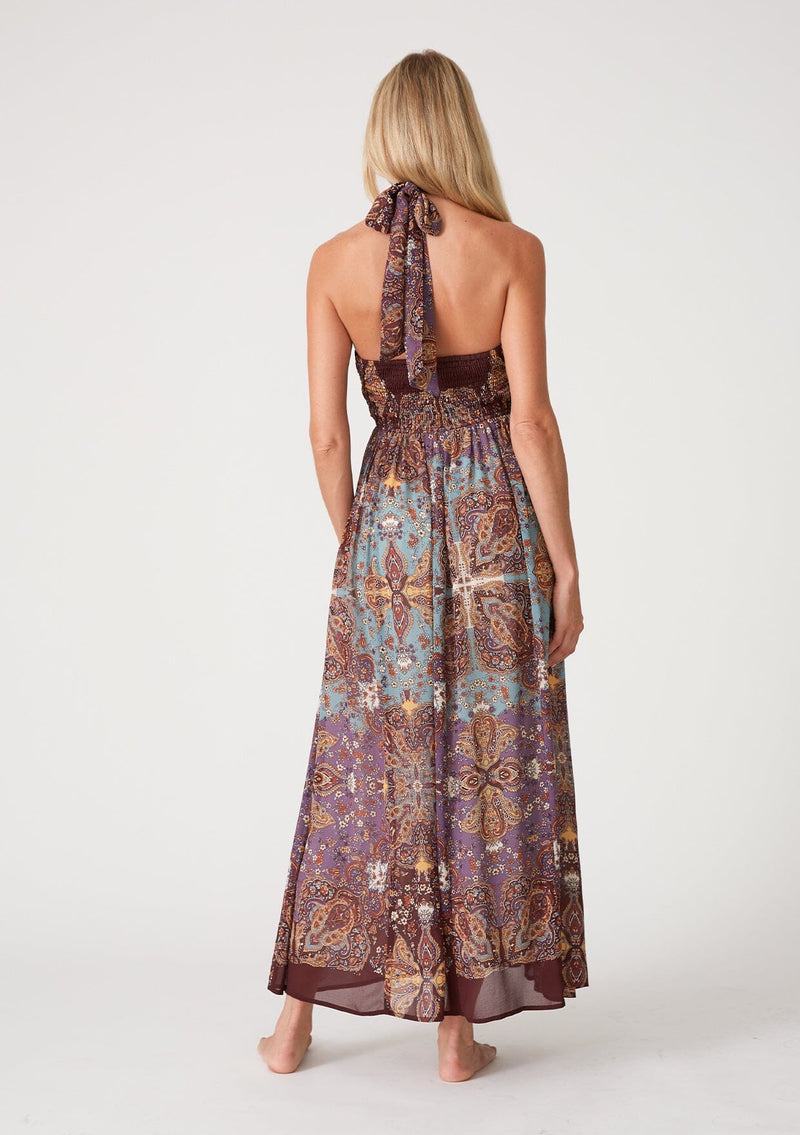[Color: Raisin/Teal] A back facing image of a blonde model wearing a bohemian halter maxi dress in a multi colored purple and teal floral print. With a tie neckline, a slim fit bodice with a smocked elastic back, and a long flowy skirt.