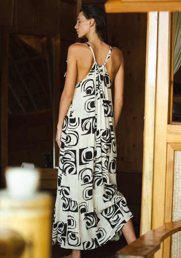 [Color: Ivory/Black] A front facing image of a brunette model wearing a flowy sleeveless bohemian maxi dress designed in an ivory and black print. With a tiered skirt and a drawstring neckline with adjustable tie.