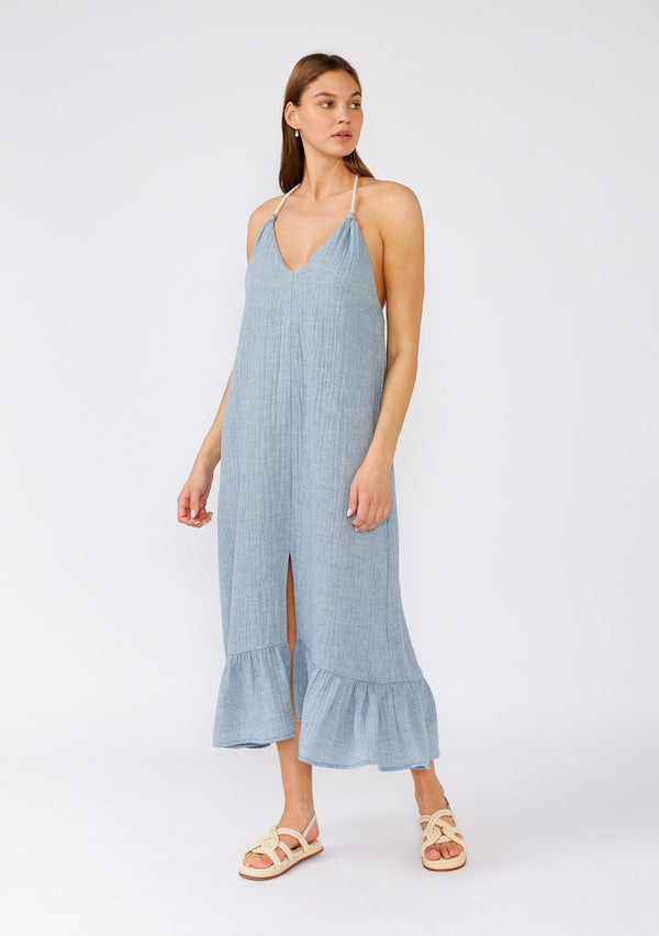 [Color: Dusty Blue] A front facing image of a brunette model wearing a dusty blue bohemian halter beach dress designed in soft cotton gauze. With a v neckline, a long tiered skirt with front slit, an adjustable rope halter tie neckline, and a t back strap detail. 