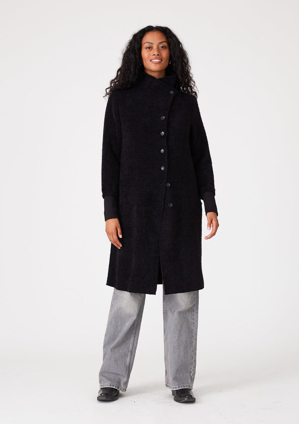 [Color: Black] A front facing image of a brunette model wearing a soft black mid length coat. With long sleeves, a button front, side pockets, and a funnel neckline.