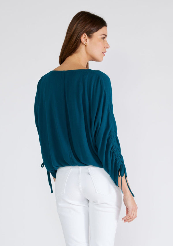 [Color: Peacock] A back facing image of a brunette model wearing a dark teal blue bohemian top. With a v neckline, a button front, an elastic waist, and three quarter length sleeves with adjustable ties. 