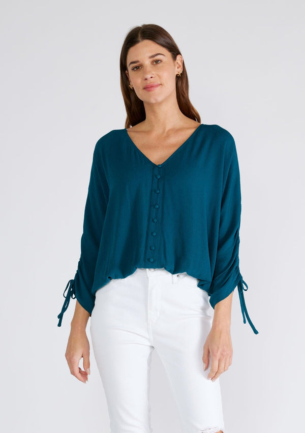 [Color: Peacock] A front facing image of a brunette model wearing a dark teal blue bohemian top. With a v neckline, a button front, an elastic waist, and three quarter length sleeves with adjustable ties. 