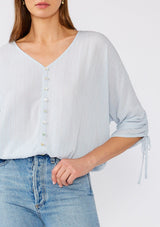 [Color: Periwinkle] A close up front facing image of a brunette model wearing a light blue bohemian top with gold metallic thread details. With three quarter length sleeves, gathered drawstring sleeve with ties, a v neckline, a button front, and an elastic waist. 