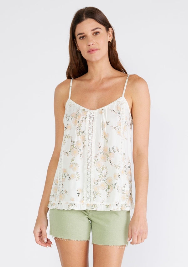 [Color: Ivory/Dusty Coral] A front facing image of a brunette model wearing a flowy bohemian camisole in an ivory and pink floral print. With adjustable spaghetti straps, a v neckline, lace trim, a ruffled hemline, and pintuck details. 