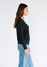 [Color: Black] A side facing image of a brunette model wearing a black soft ribbed knit pullover. With long dolman sleeves and a wide boat neck that can be worn on or off the shoulder. 