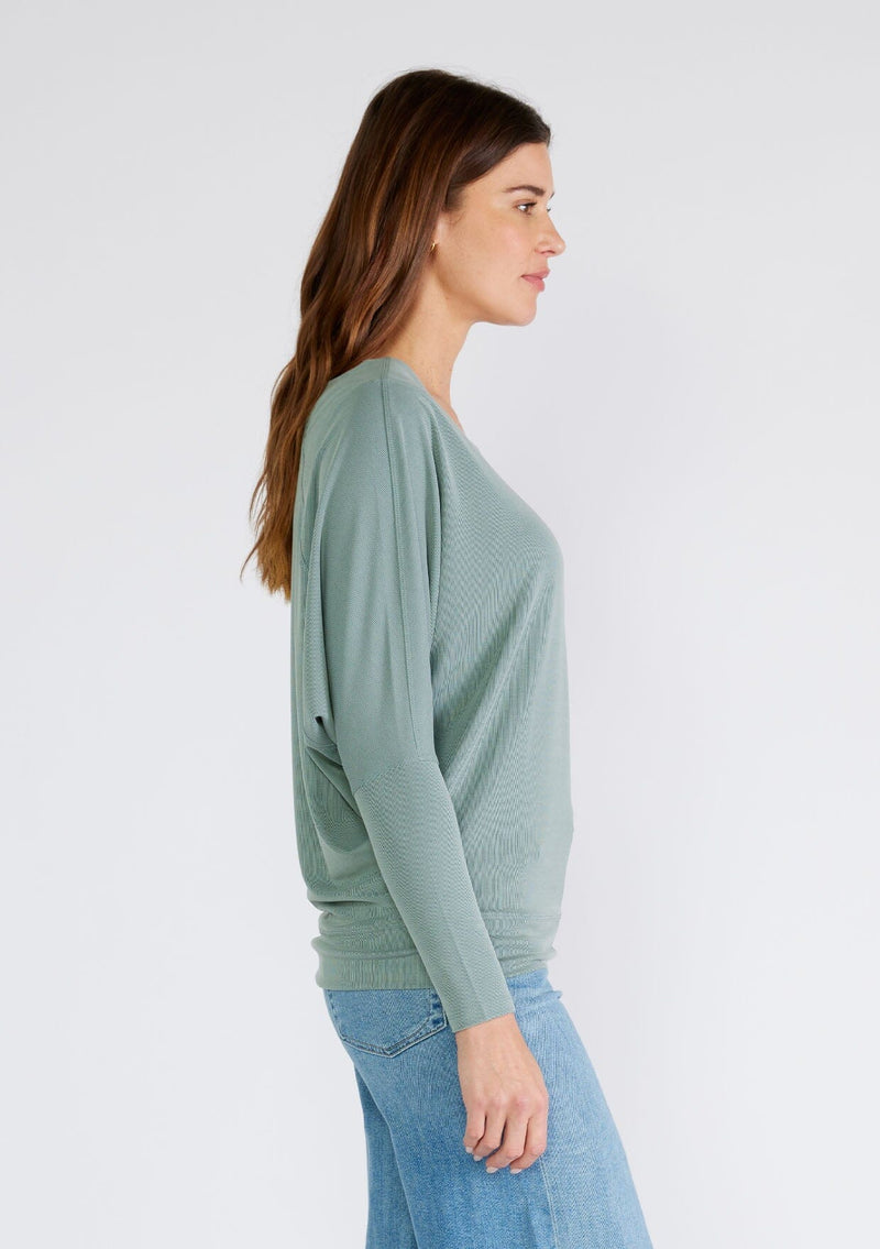 [Color: Slate Green] A side facing image of a brunette model wearing a seafoam green soft ribbed knit pullover. With long dolman sleeves and a wide boat neck that can be worn on or off the shoulder. 