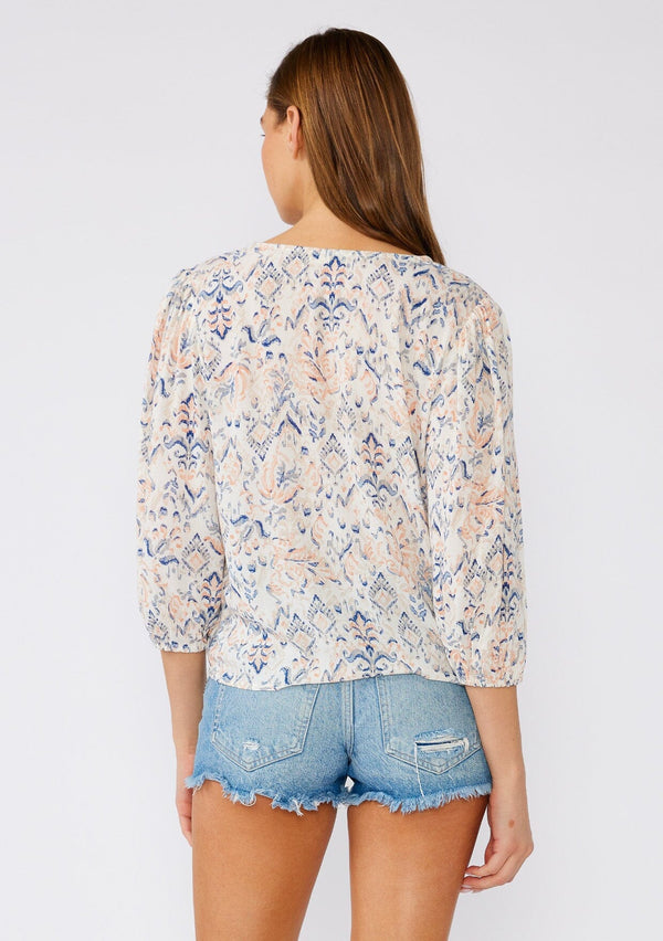 [Color: Cream/Dusty Blue] A back facing image of a brunette model wearing a lightweight bohemian blouse designed in an off white and blue print. With a v neckline, a tie waist detail, and three quarter length sleeves. 