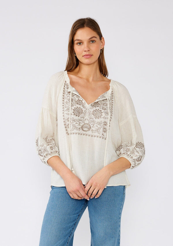[Color: Natural/Taupe] A front facing image of a brunette model wearing an ivory flowy bohemian peasant top with embroidered detail and tassel ties.