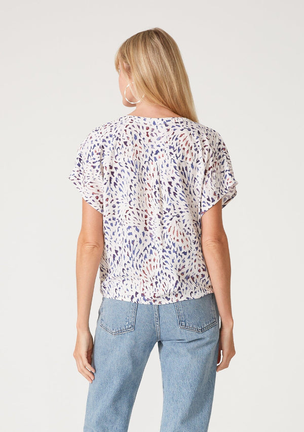[Color: Natural/Ink] A back facing image of a blonde model wearing a casual resort top in a multi colored abstract print. With short sleeves, a loop button front, a v neckline, and a tie front waist detail. 