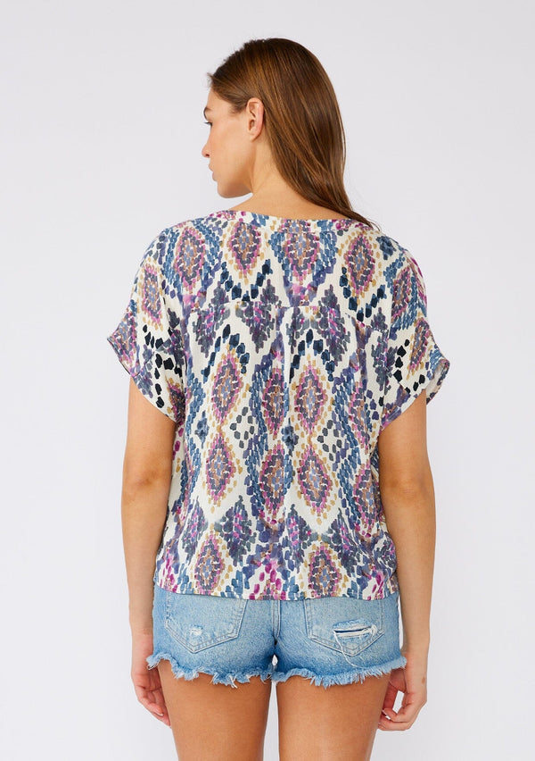[Color: Natural/Dusty Blue] A back facing image of a brunette model wearing a timeless bohemian top designed in a blue abstract bohemian print. With short sleeves, a v neckline, a self covered button front, and a tie waist detail. 