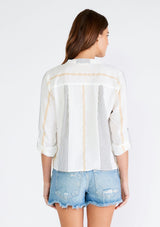 [Color: White/Gold] A back facing image of a brunette model wearing a white cotton top in a textured black embroidered stripe and light catching gold stripe detail. With long rolled sleeves and a button tab closure, a v neckline, a concealed button front, and a tie waist detail. 