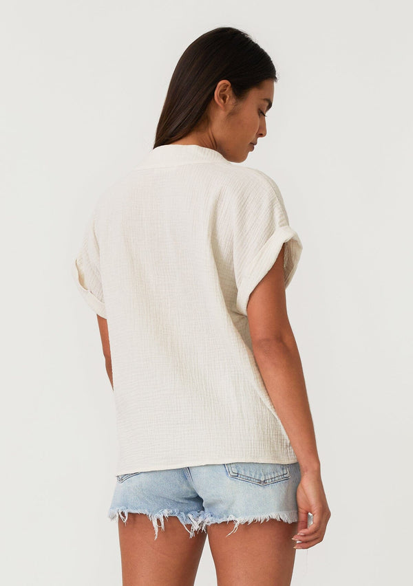 [Color: Cream] A back facing image of a brunette model wearing an off white cotton gauze tee. With short cuffed sleeves, a v neckline, and an oversized relaxed fit. 