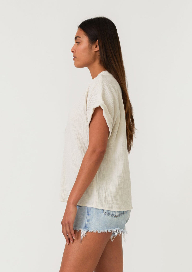[Color: Cream] A side facing image of a brunette model wearing an off white cotton gauze tee. With short cuffed sleeves, a v neckline, and an oversized relaxed fit. 