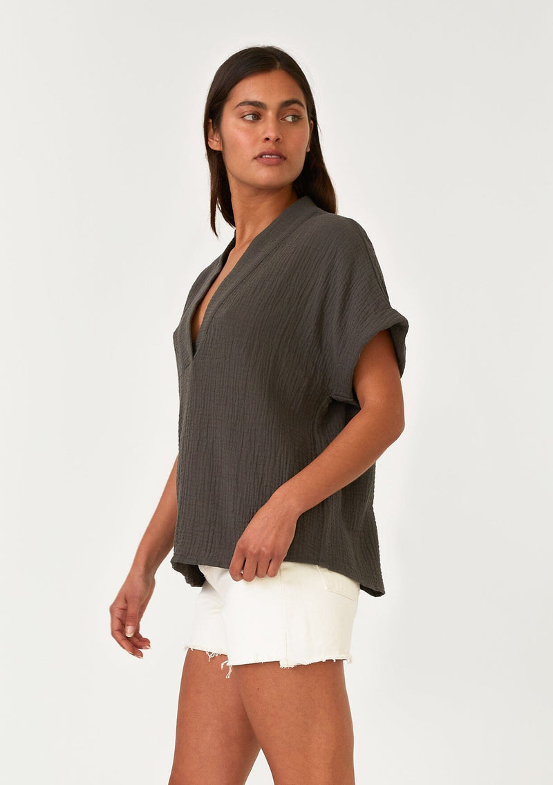 [Color: Lagoon] A side facing image of a brunette model wearing an olive green cotton gauze tee. With short cuffed sleeves, a v neckline, and an oversized relaxed fit. 