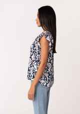 [Color: Navy/Natural] A side facing image of a brunette model wearing a blue floral summer blouse. With short flutter sleeves, a split v neckline with double tassel ties, and a relaxed fit. 
