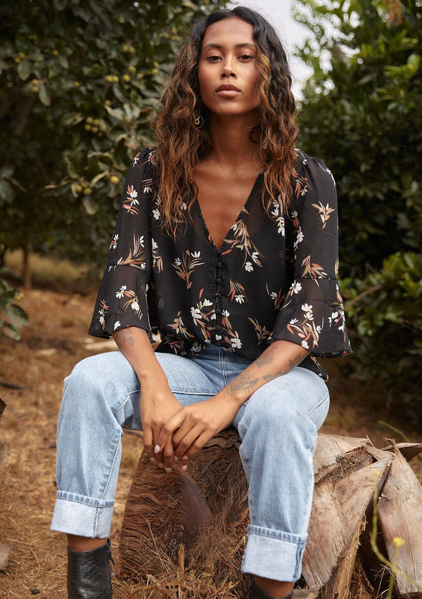 [Color: Black/Peach] A front facing image of a black model sitting outside wearing a delicate chiffon bohemian blouse in a black and peach floral print. With half length flutter sleeves, a v neckline, and a self covered loop button front.