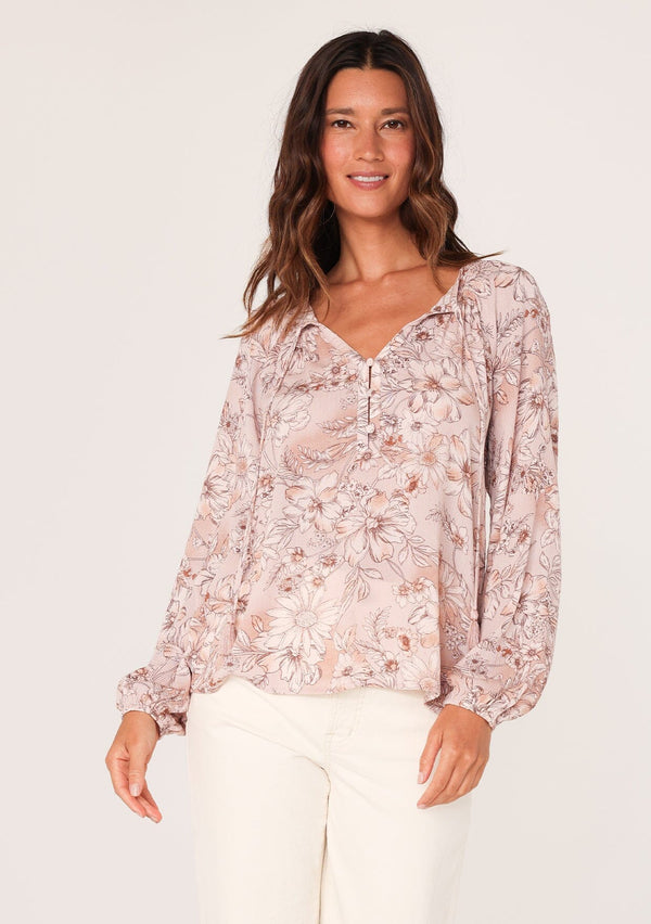 [Color: Dusty Rose/Light Brown] A front facing image of a brunette model wearing a boho blouse in a pink floral print. With long sleeves, a self covered button front, a split v neckline with tassel ties, and smocked details along the neckline. 