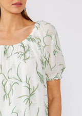 [Color: Off White/Green] A close up front facing image of a brunette model wearing a bohemian cotton top in a white and green embroidery. With short puff sleeves, a ruffled elastic cuff, an elastic scoop neckline, and a relaxed, flowy fit. 