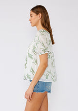 [Color: Off White/Green] A side facing image of a brunette model wearing a bohemian cotton top in a white and green embroidery. With short puff sleeves, a ruffled elastic cuff, an elastic scoop neckline, and a relaxed, flowy fit. 