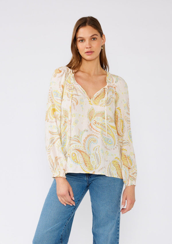 [Color: Dusty Peach/Caramel] A front facing image of a brunette model wearing a bohemian blouse in a pink and yellow paisley print. With gold lurex details, long raglan sleeves, a split v neckline with optional hook and eye closure, neck ties, and smocked details at the neckline. 