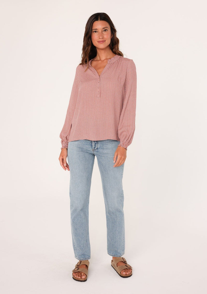 [Color: Mauve] A full body front facing image of a brunette model wearing a bohemian fall blouse in a mauve pink dot print. With long sleeves, a ruffled elastic wrist cuff, a self covered button front, a ruffled neckline, and a split v neckline.