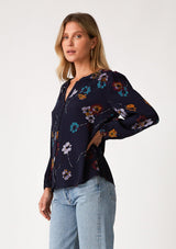 [Color: Navy/Mustard] A side facing image of a blonde model wearing a bohemian fall blouse in a blue floral print. With long sleeves, a v neckline, a self covered button front, and a relaxed fit. 