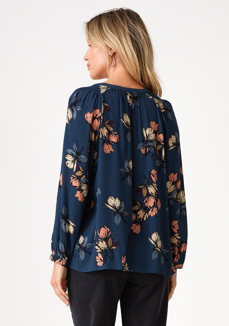 [Color: Teal/Dusty Blush] A back facing image of a blonde model wearing a bohemian fall blouse in a teal blue floral print. With voluminous long raglan sleeves, a v neckline, a smocked shoulder detail, and a self covered button front. 
