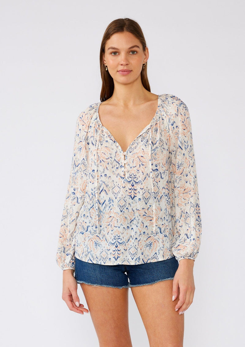 [Color: Cream/Dusty Blue] A front facing image of a brunette model wearing a lightweight bohemian blouse in an abstract white and blue print. With long raglan sleeves, a v neckline with ties, a self covered button front, and a relaxed fit. 