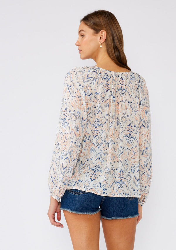 [Color: Cream/Dusty Blue] A back facing image of a brunette model wearing a lightweight bohemian blouse in an abstract white and blue print. With long raglan sleeves, a v neckline with ties, a self covered button front, and a relaxed fit. 