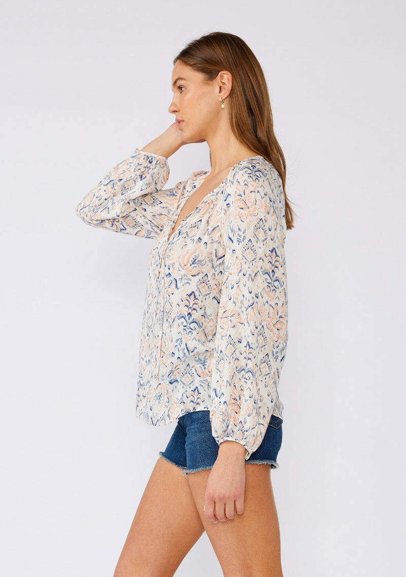 [Color: Cream/Dusty Blue] A side facing image of a brunette model wearing a lightweight bohemian blouse in an abstract white and blue print. With long raglan sleeves, a v neckline with ties, a self covered button front, and a relaxed fit. 
