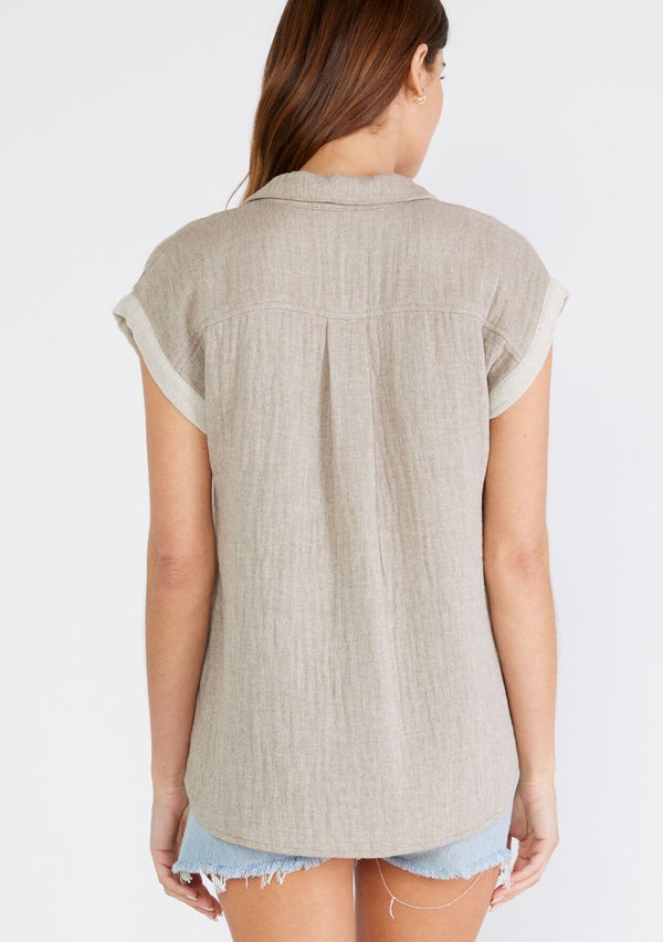 [Color: Taupe] A back facing image of a brunette model wearing a light brown short sleeve shirt crafted from soft cotton gauze. With a button up front, a collared neckline, a front patch pocket, and short contrast cap sleeves. 