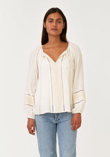 [Color: Cream/Natural] A front facing image of a brunette model wearing a cream bohemian fall blouse with embroidered details and sparkly metallic thread. With long raglan sleeves, a split v neckline with tassel ties, lattice trim, and a relaxed fit.