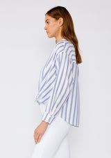 [Color: White/Blue] A side facing image of a brunette model wearing a relaxed spring shirt in a white and blue stripe and a metallic gold thread detail. With long sleeves, a dropped shoulder, a v neckline, a collared neckline, a button front, and a knot waist detail. 