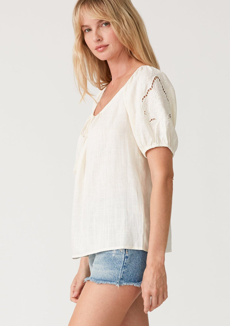 [Color: Vanilla] A side facing image of a blonde model wearing an off white bohemian resort top with embroidered details. With short puff sleeves and a wide scooped neckline with a drawstring and tassel ties. 