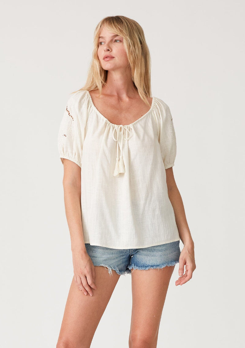 [Color: Vanilla] A front facing image of a blonde model wearing an off white bohemian resort top with embroidered details. With short puff sleeves and a wide scooped neckline with a drawstring and tassel ties. 