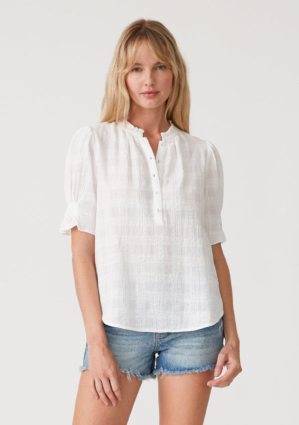 [Color: White] A front facing image of a blonde model wearing a white bohemian cotton blend blouse in a textured gingham. With short puff sleeves, a button front, a high ruffled neckline, and smocked elastic details at the sleeve.