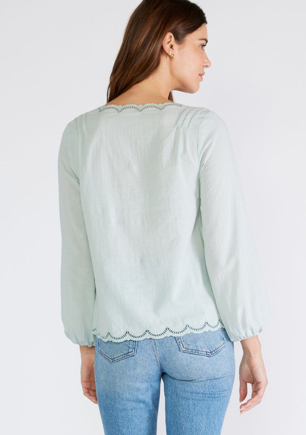 [Color: Dusty Seafoam] A back facing image of a brunette model wearing a feminine spring blouse in dusty seafoam. With scalloped embroidered lace trim, a button front, a v neckline, and long sleeves with elastic wrist cuffs. 