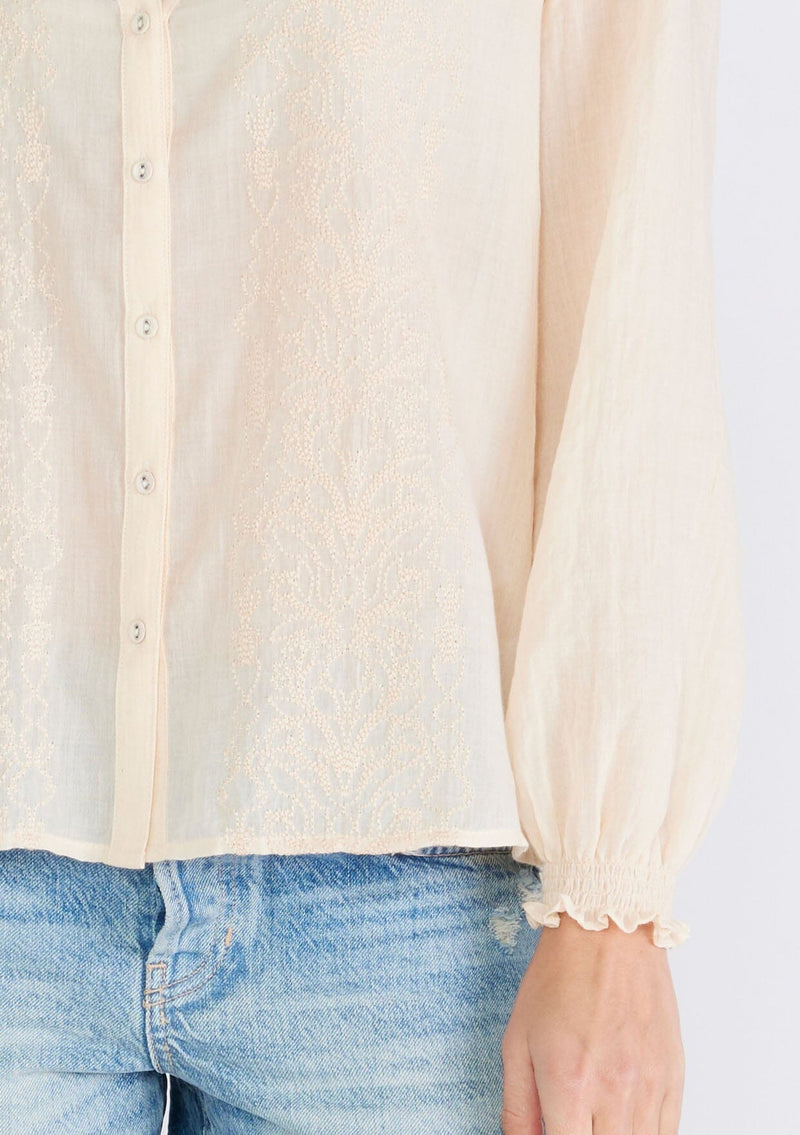 [Color: Light Peach] A close up front facing image of a brunette model wearing a sheer bohemian blouse in light peach pink. With long sleeves, smocked elastic wrist cuffs, a self covered button front, a split v neckline, and embroidered details along the front. 