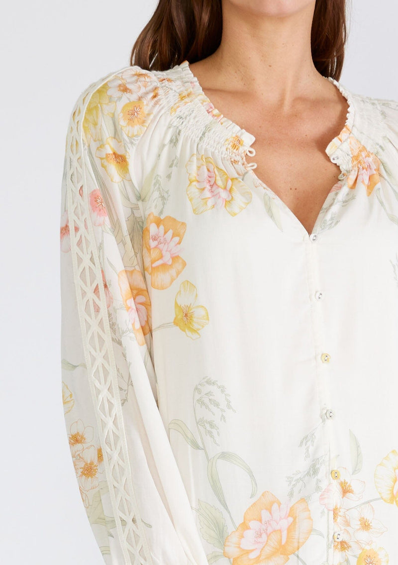 [Color: Vanilla/Coral] A close up front facing image of a brunette model wearing a beautiful bohemian chic spring blouse in an ivory and coral floral print. With long raglan sleeves, a smocked round neckline, a self covered button front, and a front keyhole. 