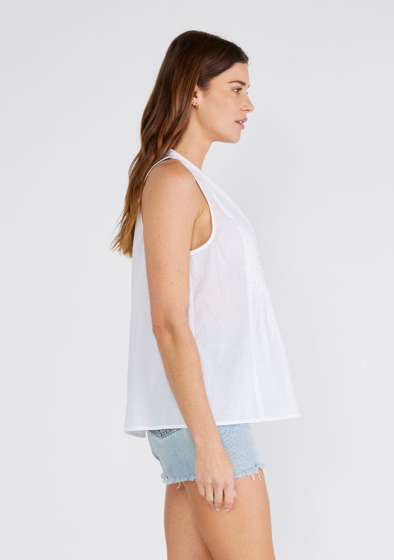 [Color: Chalk] A side facing image of a brunette model wearing a bohemian cotton tank top in a white. With a v neckline, a loose, tent silhouette, and pleated details along the front.
