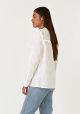 [Color: White] A side facing image of a brunette model wearing a white bohemian button front blouse with a ruffled round neckline, a self covered button front, long sleeves, and an eyelet yoke detail. 