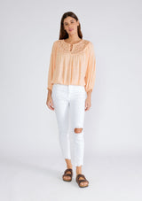 [Color: Mellow Peach] A full body front facing image of a brunette model wearing a peach bohemian blouse with long sleeves, a ruffled elastic wrist cuff, a billowy silhouette, an elastic hemline, and a sheer crochet yoke detail. 