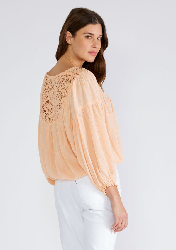 [Color: Mellow Peach] A back facing image of a brunette model wearing a peach bohemian blouse with long sleeves, a ruffled elastic wrist cuff, a billowy silhouette, an elastic hemline, and a sheer crochet yoke detail. 