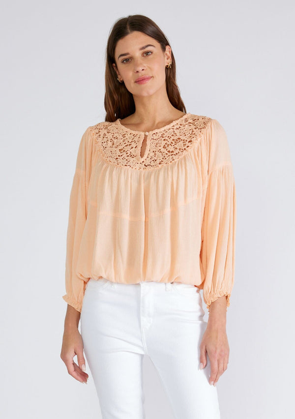[Color: Mellow Peach] A front facing image of a brunette model wearing a peach bohemian blouse with long sleeves, a ruffled elastic wrist cuff, a billowy silhouette, an elastic hemline, and a sheer crochet yoke detail. 