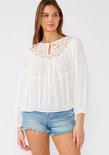 [Color: White] A half body front facing image of a brunette model wearing a white bohemian blouse with long sleeves, a ruffled elastic wrist cuff, a billowy silhouette, an elastic hemline, and a sheer crochet yoke detail. 