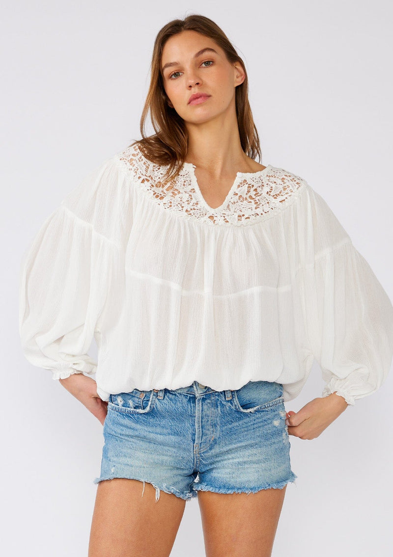 [Color: White] A front facing image of a brunette model wearing a white bohemian blouse with long sleeves, a ruffled elastic wrist cuff, a billowy silhouette, an elastic hemline, and a sheer crochet yoke detail. 