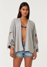 [Color: Silver/Teal] A half body front facing image of a brunette model wearing a bohemian kimono top with a silver grey exterior and teal blue interior. With voluminous long sleeves, floral embroidery, an open front, and tie wrist cuffs. 