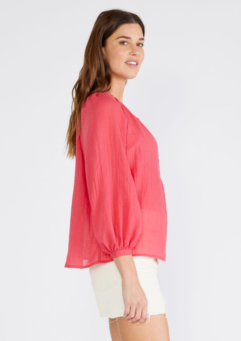[Color: Coral] A side facing image of a brunette model wearing a red sheer bohemian blouse with long raglan sleeves, a round neckline, a button front, and a relaxed fit. 